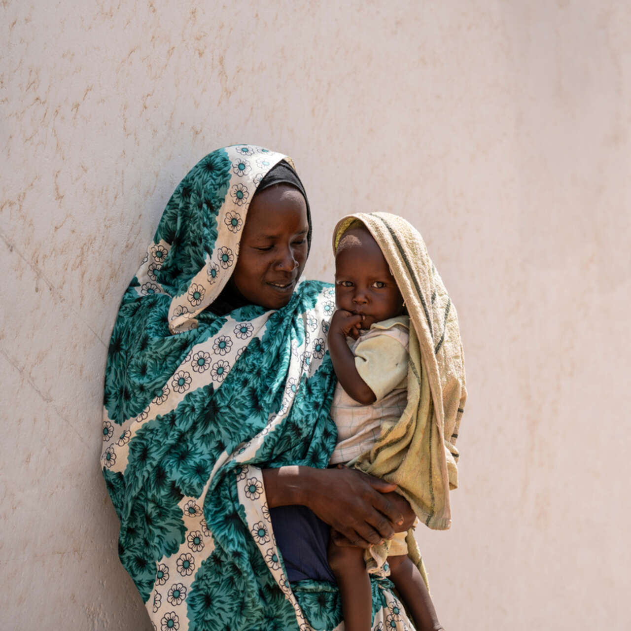 En mamma som flytt från Sudan står lutad mot en vägg och håller sin son i famnen.